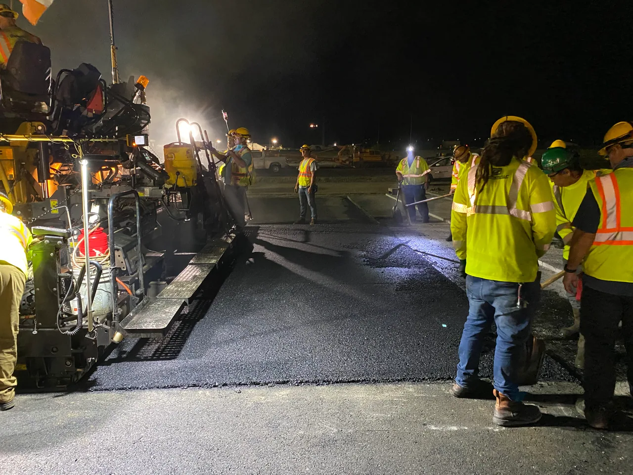 Crew members finishing fresh asphalt beside a runway paver at night.