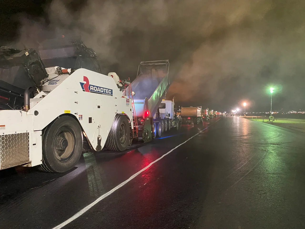 Material transfer equipment and trucks lined up for nighttime runway paving.