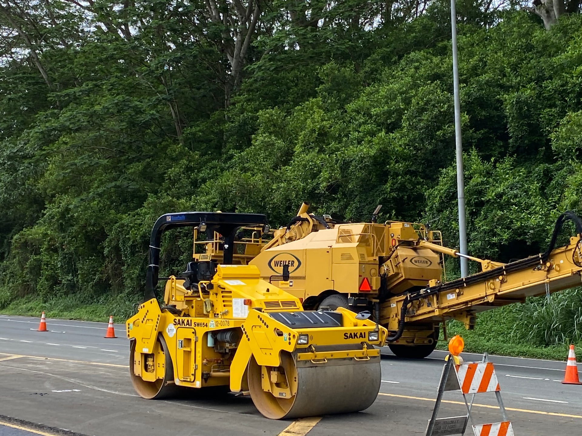 A roller and paving equipment positioned beside a coned-off roadway.